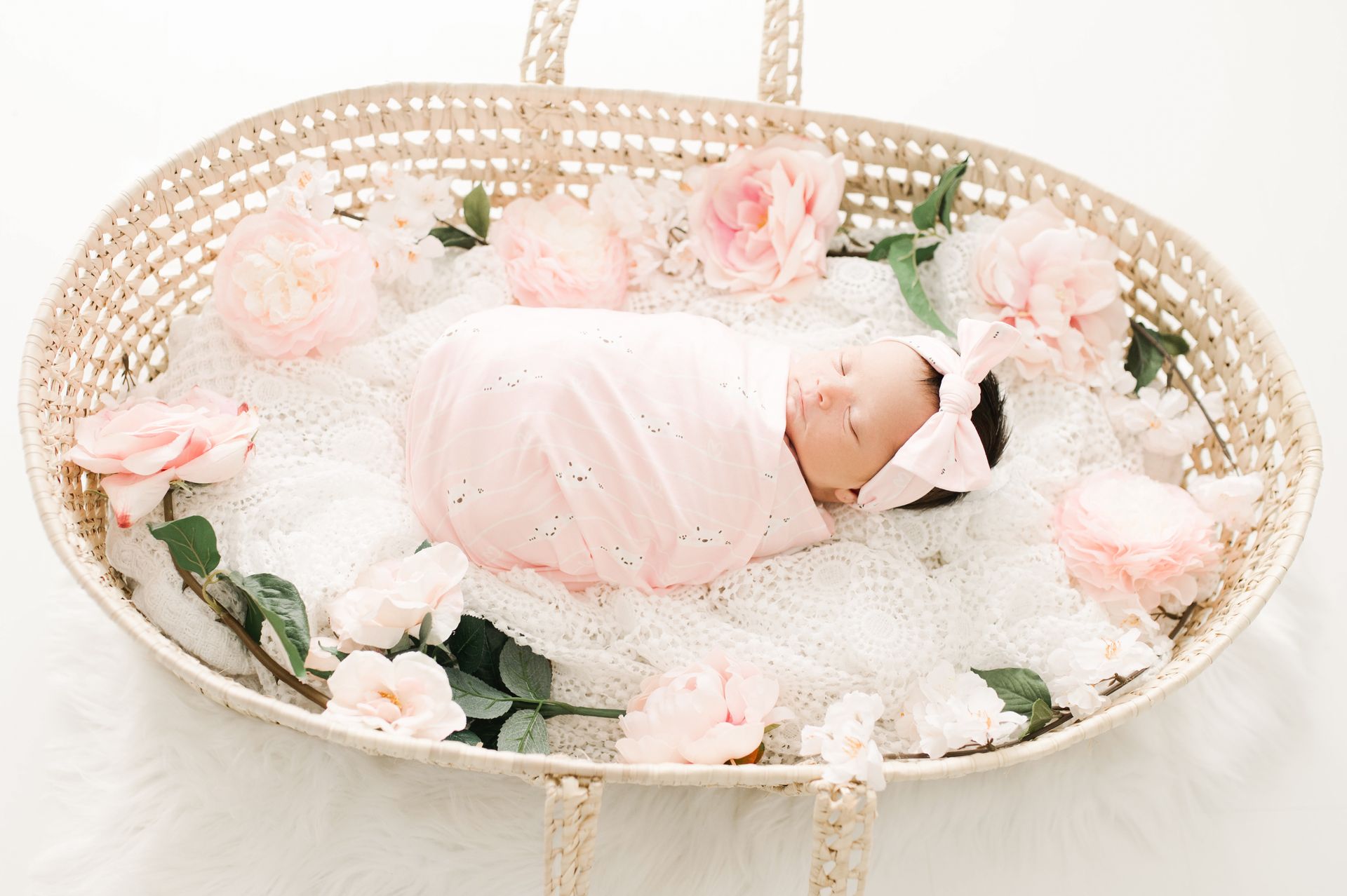 Baby in a pink swaddle and bow in a Moses basket beside roses