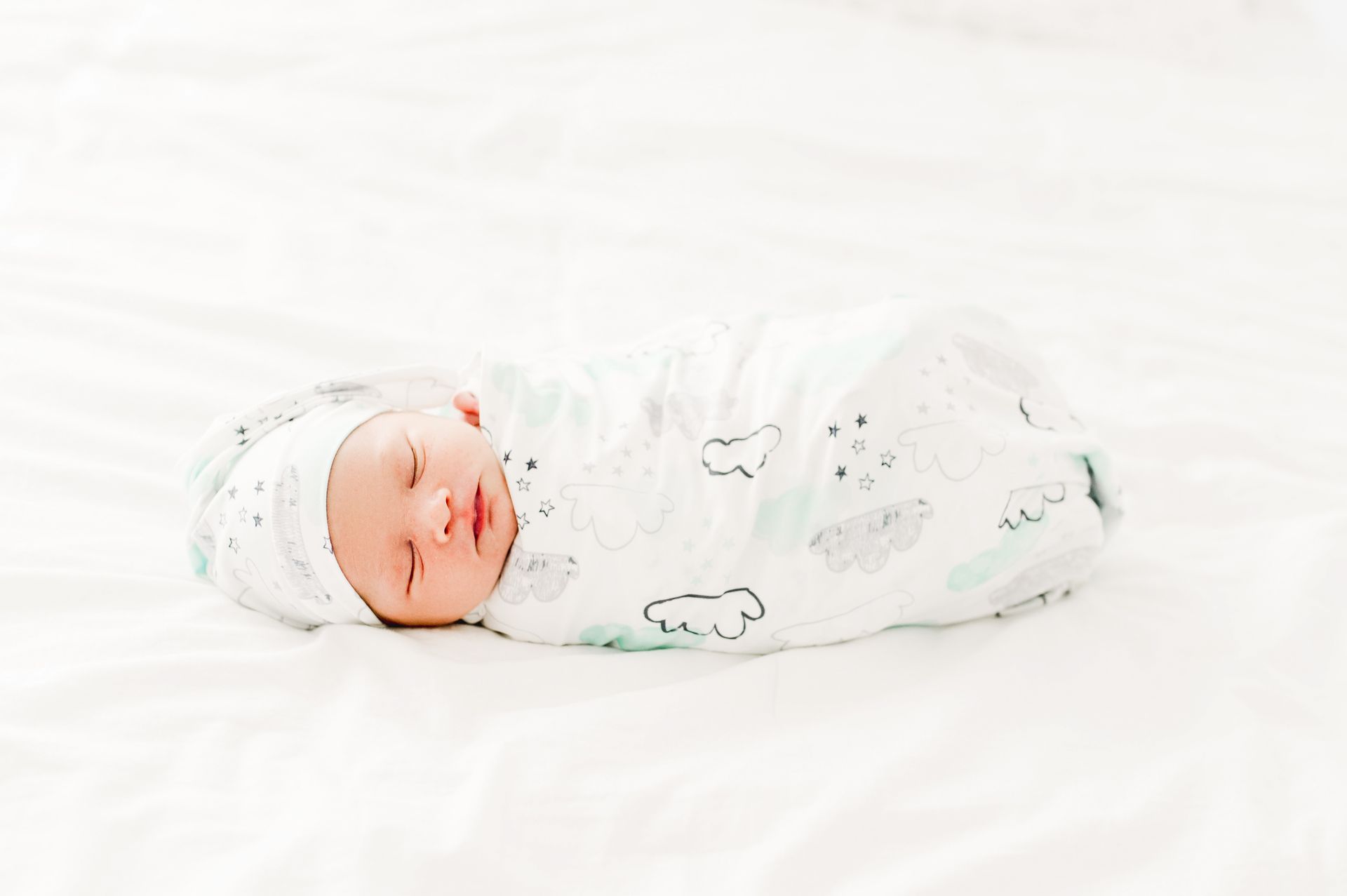 Baby in a cloud-and-star-print swaddle and hat on a white bed